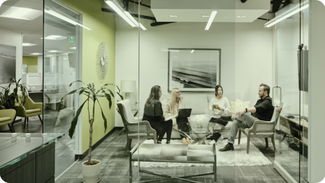 Four people in a conference room with glass walls, sitting at a table and participating in a discussion.